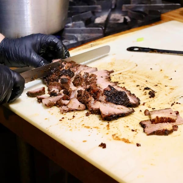 Chef in black gloves slicing smoked brisket on a white cutting board, revealing juicy pink meat.