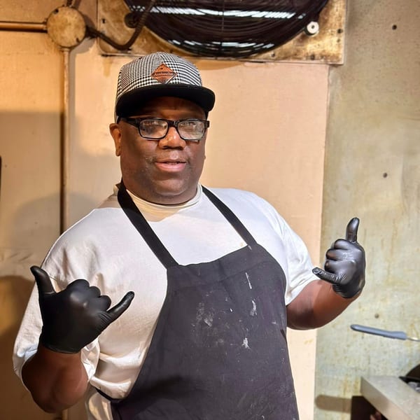 Blind Raven BBQ chef in a houndstooth hat and black apron, giving shaka signs.