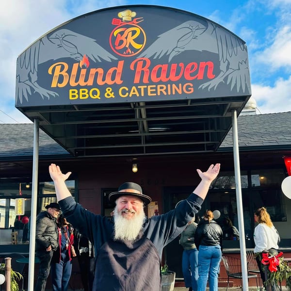 A man with a long white beard and hat stands with arms raised in front of the Blind Raven BBQ sign.