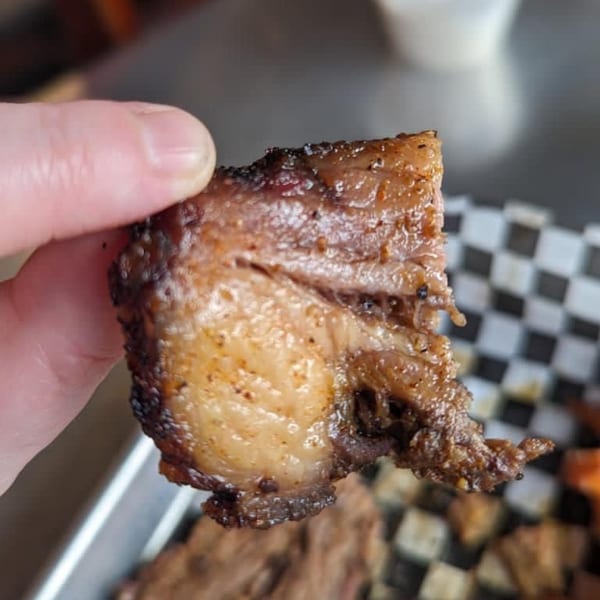 A hand holds up a piece of Blind Raven BBQ brisket, showing its smoky bark and juicy interior.