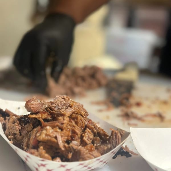 Close-up of Blind Raven BBQ's pulled pork in a paper boat, with a gloved hand in the background.