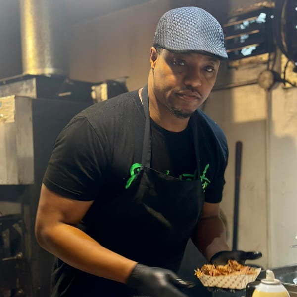 A chef in a black apron and cap holds a tray of pulled pork in a smoky kitchen.